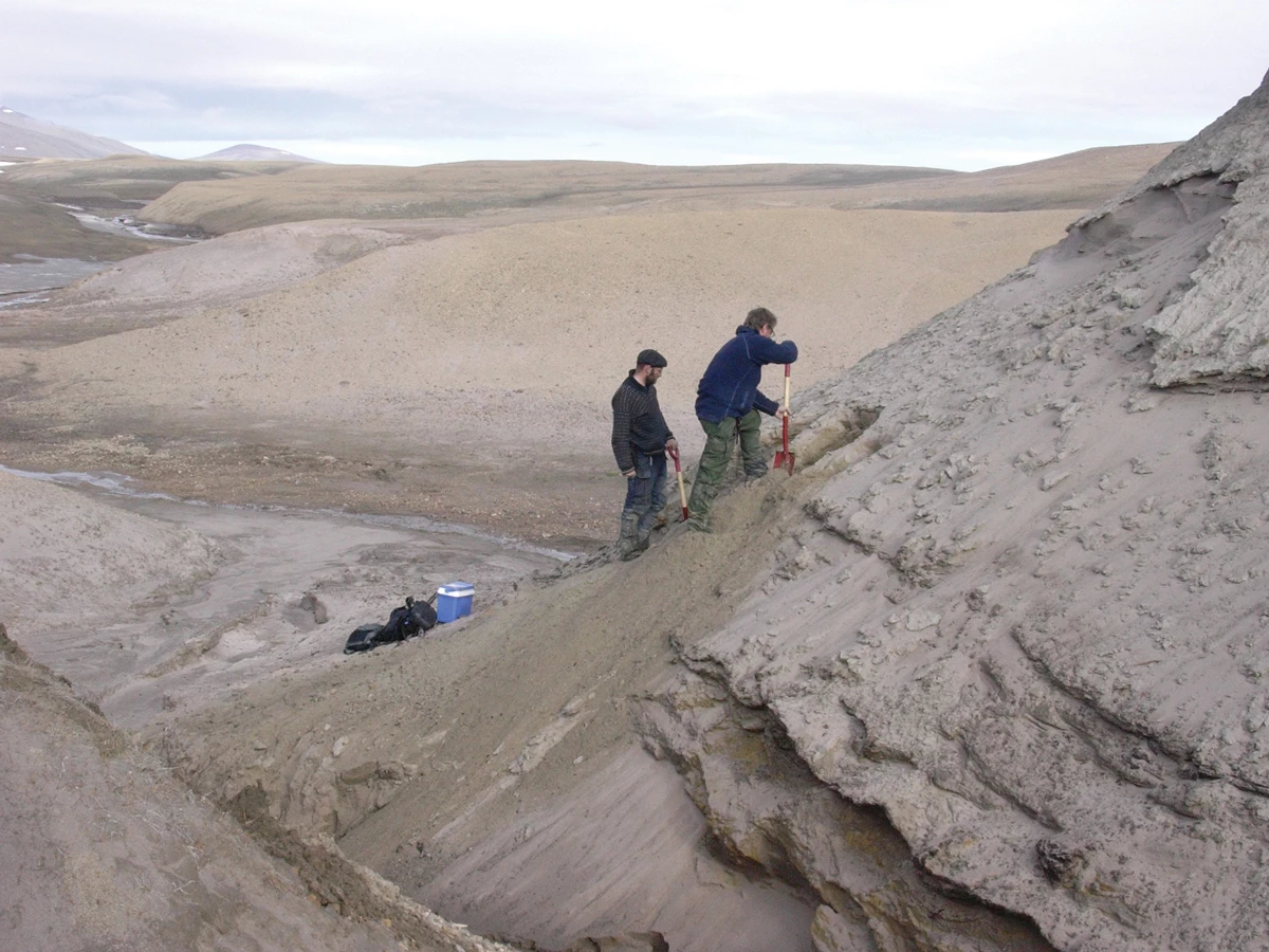 Two warmly dressed men digging on a sandy slope against a barren landscape.