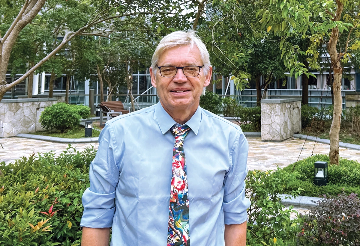 A man with white hair stands in front of a planted border. He wears glasses, a light-blue shirt, and a colorful tie. His hands are in his trouser pockets.