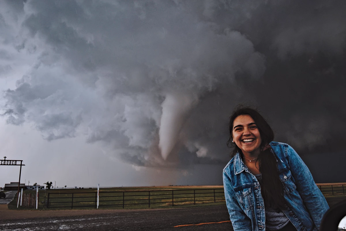 Ariana Castillo stands in front of an open field. Behind her, a tornado is forming.