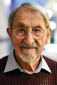 Martin Karplus, a man with circular glasses, short gray hair, and a gray goatee, looks at the viewer with a slight smile on his face.