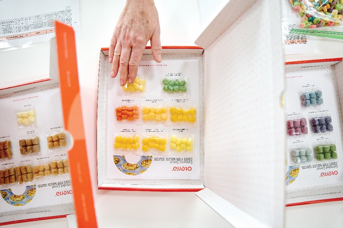 A hand points at a box containing multicolored cereal puffs. Two other boxes with cereal puffs are on either side.