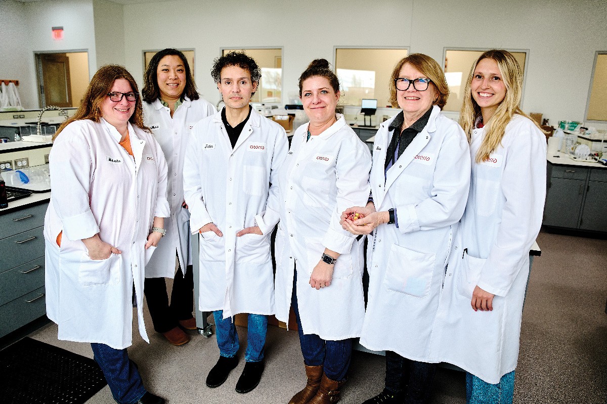 Six scientists in white lab coats stand next to each other in a laboratory. 