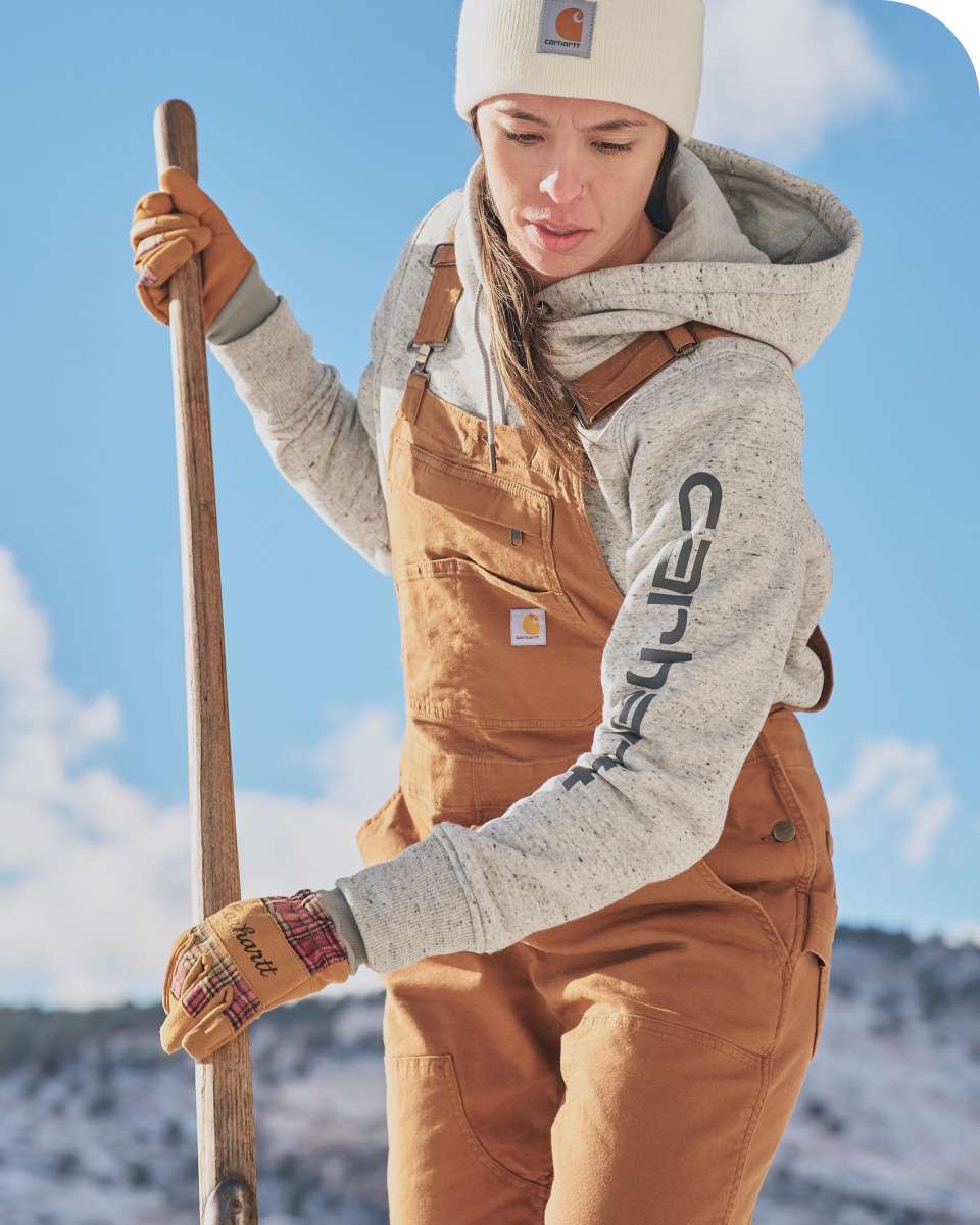 woman wearing a gray hoodie, brown overalls, a white beanie, and work gloves is shoveling outdoors in a snowy landscape. She has a focused expression, and the Carhartt logo is visible on her clothing