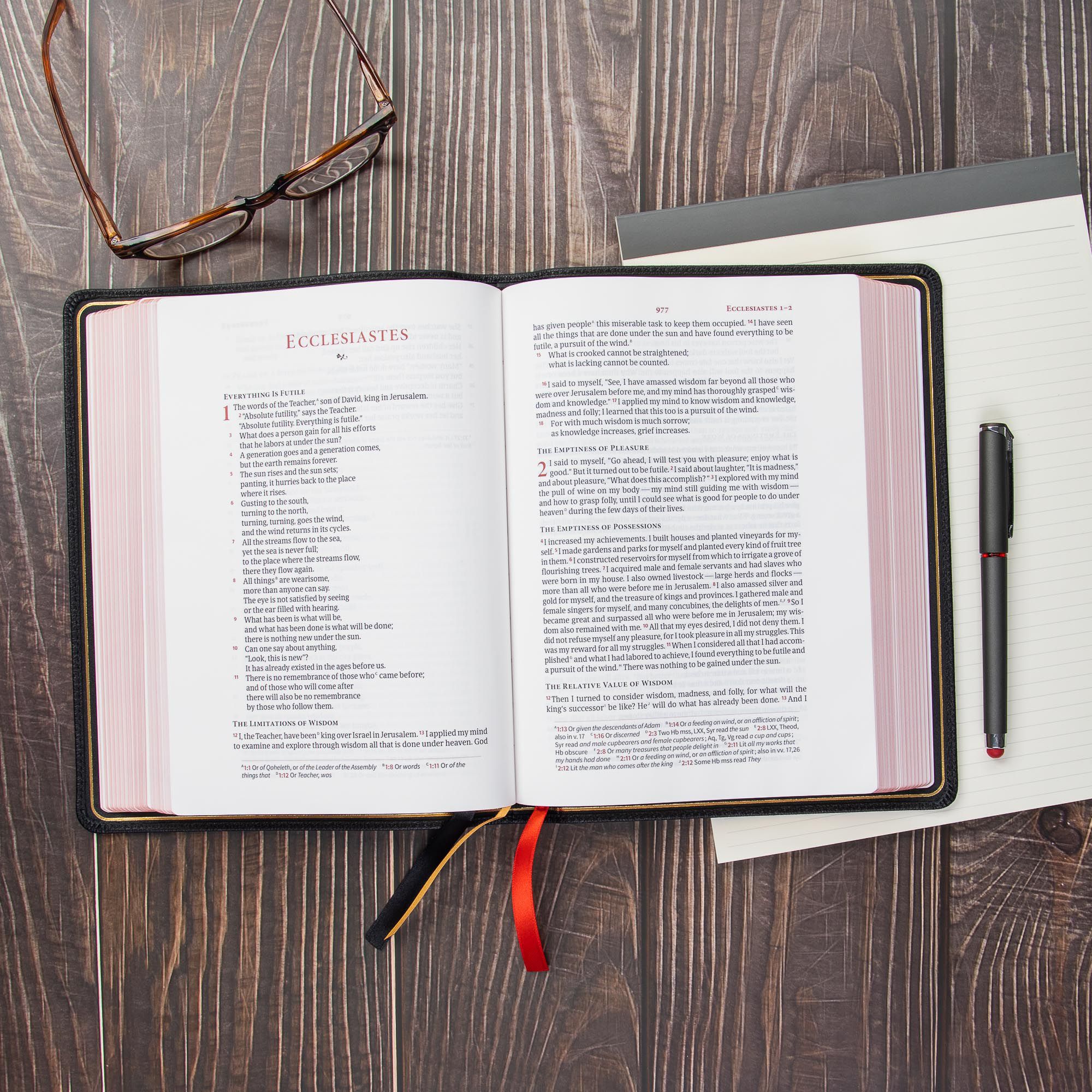 Open Bible on a wooden desk with glasses and a notebook.