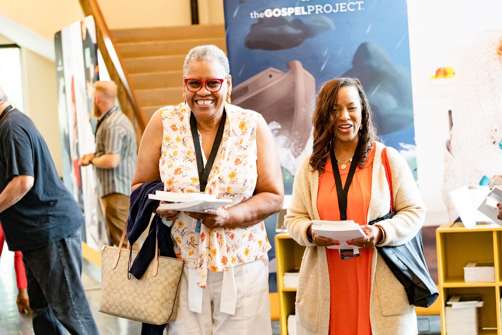 Two women smiling and holding papers at an event, with banners for The Gospel Project in the background