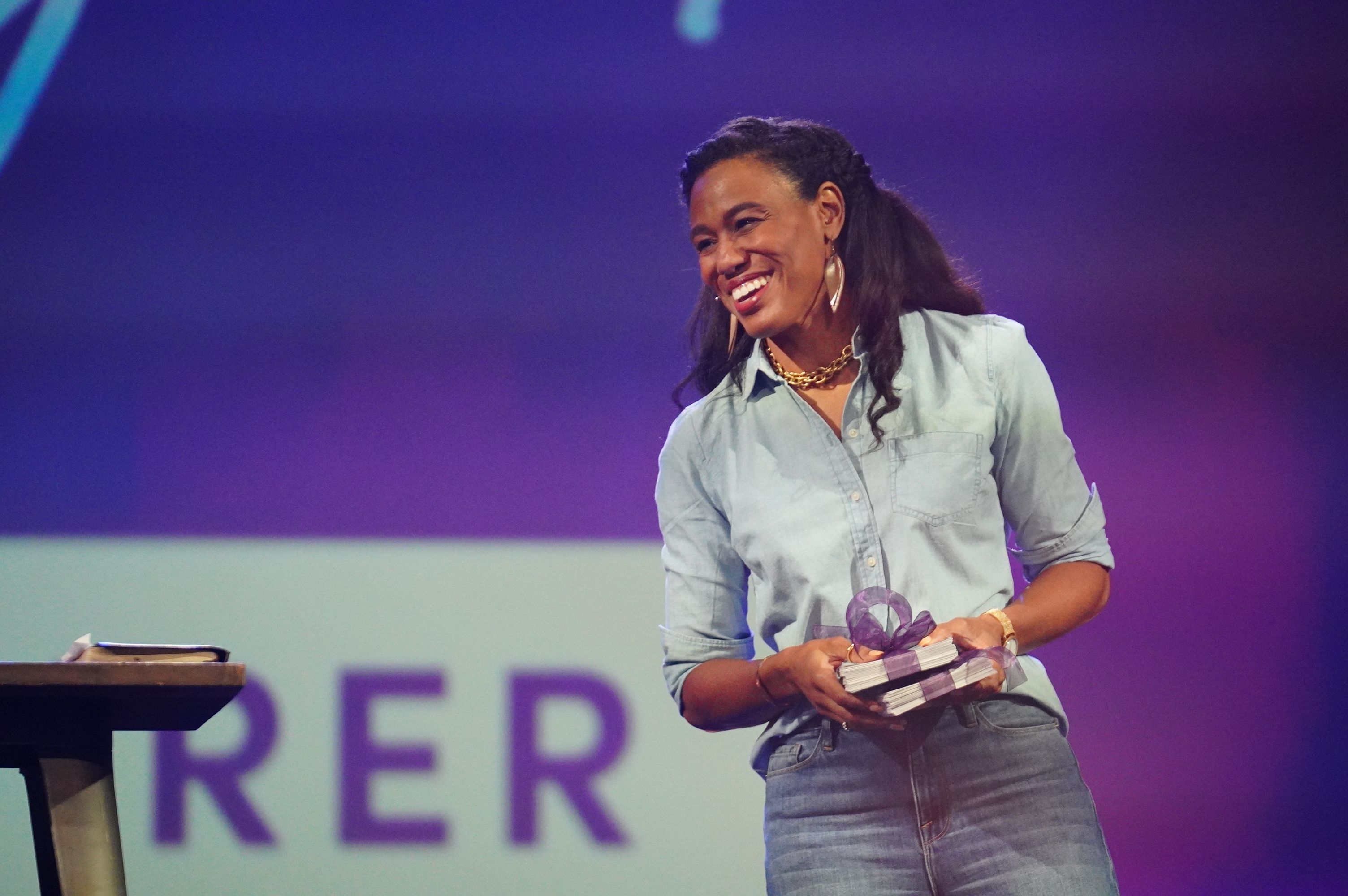 Priscilla Shirer smiling and holding gifts while standing on stage with a purple background