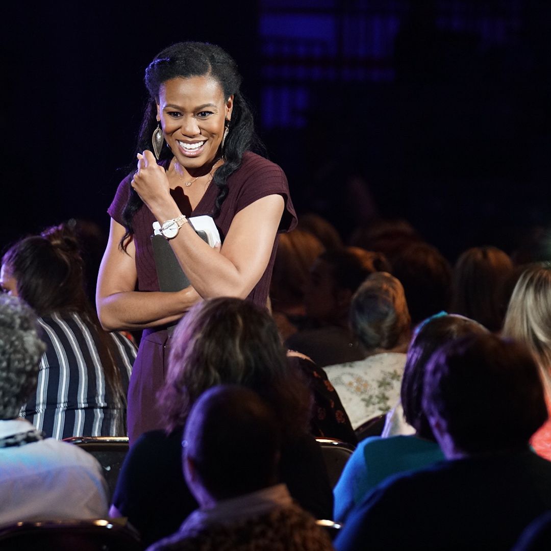 Priscilla Shirer speaking on stage with a book in hand, smiling and wearing a purple outfit