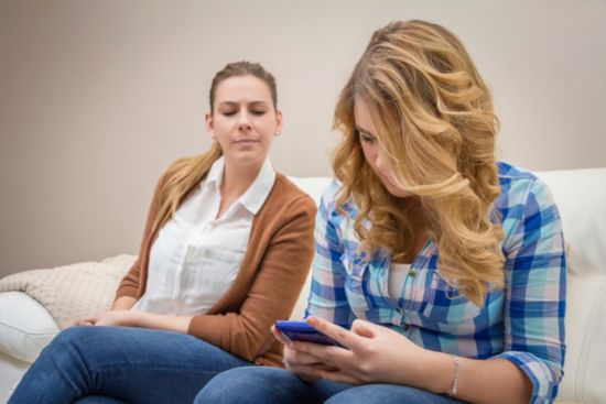 Daughter looking at smartphone sitting on couch, mom looking over her shoulder.