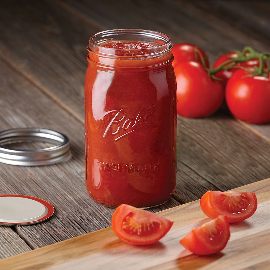 Ball wide mouth jar filled with tomato sauce on a table setting with fresh tomatoes in background