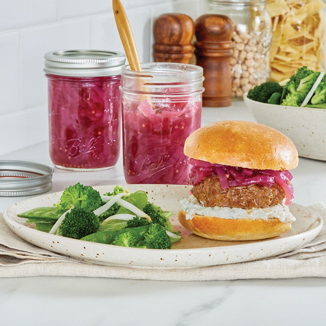Jars with pickled red onions behind a burger garnished with red onions