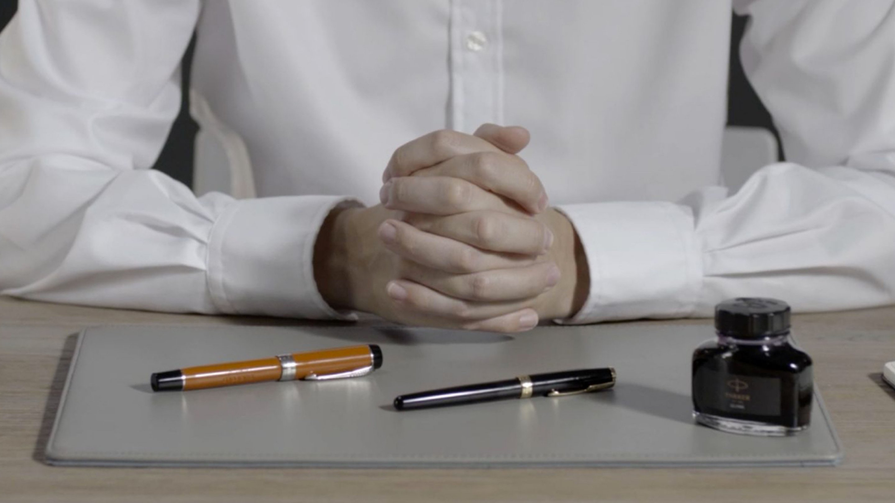 A person with hands folded sitting at a desk with two capped pens and a bottle of ink.