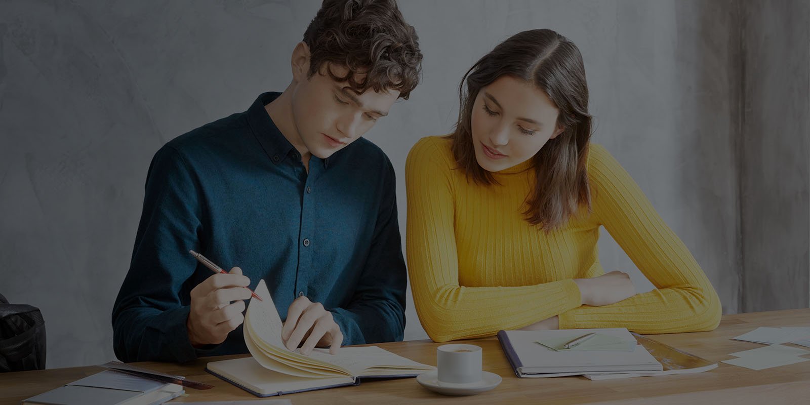 A young man holding a pen and notebook while sitting next to a young woman at a desk.