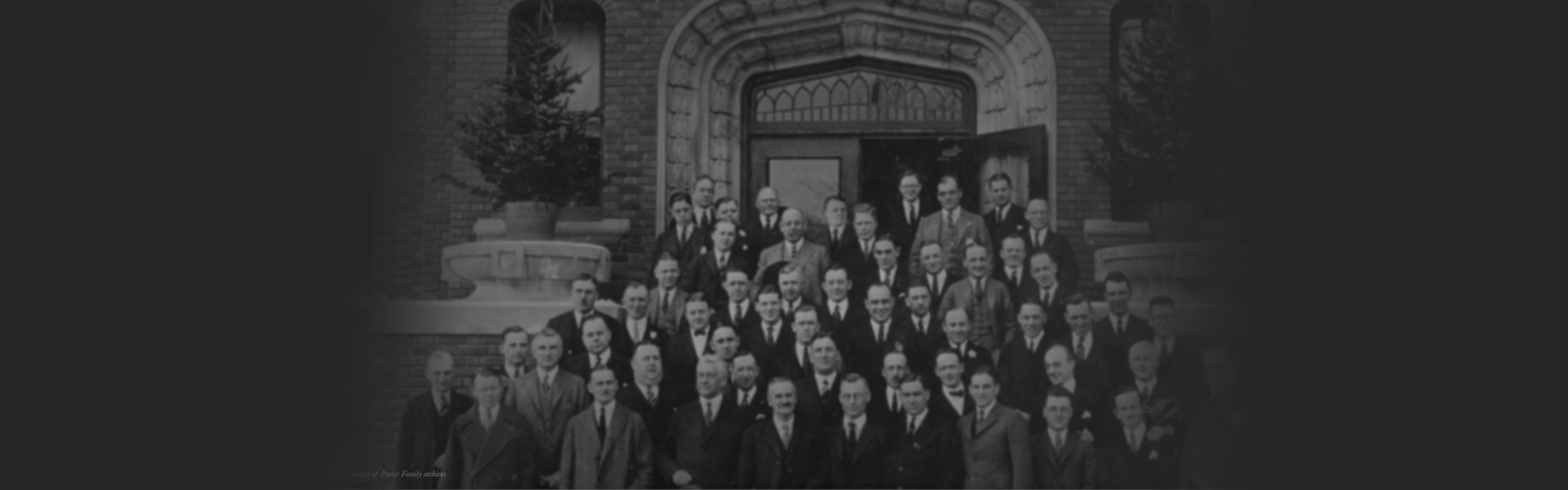 A vintage photograph of many men in suits in front of a building.