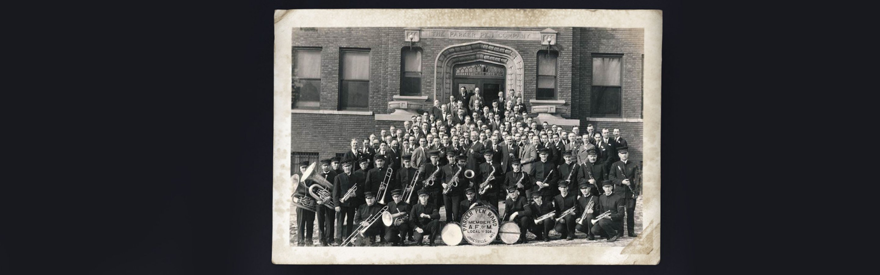 A vintage photograph of the Parker Pen marching band and men in suits in from of the Parker Pen factory.
