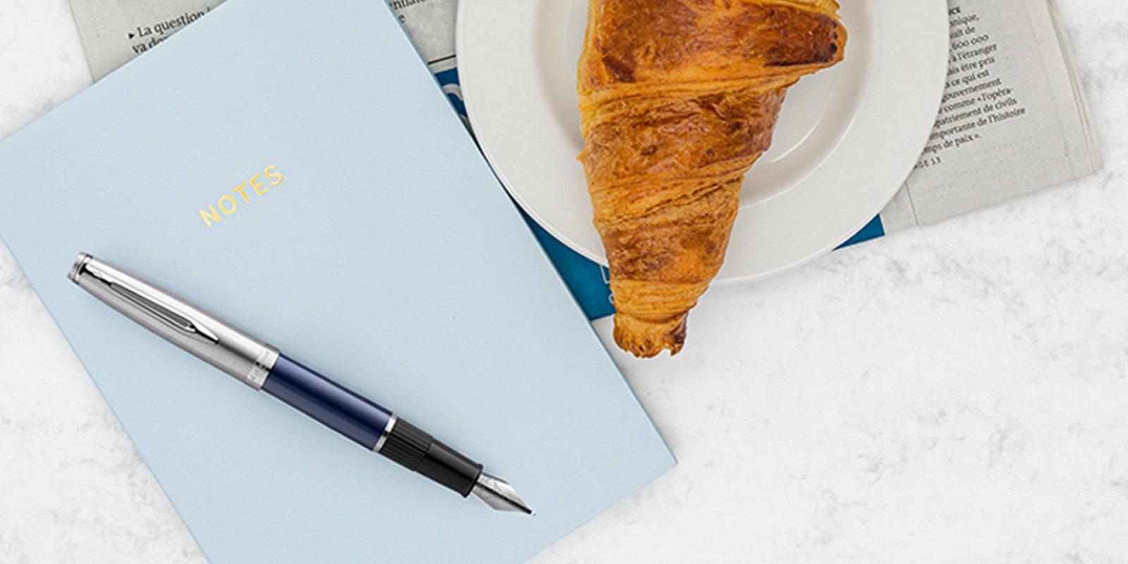 An Embleme fountain pen with chrome trim laid on a notebook next to a croissant, cup of coffee and newspaper.