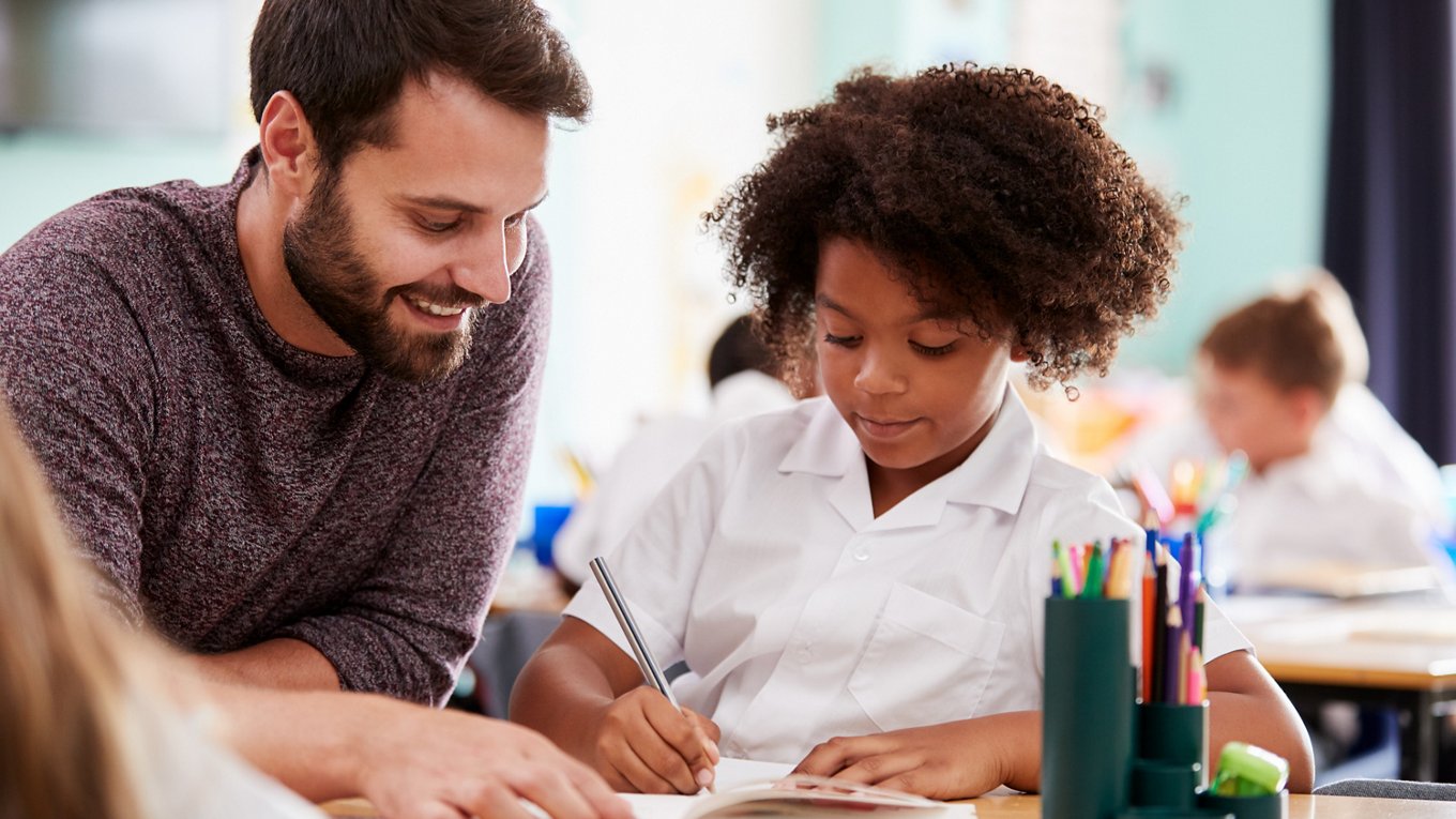 An male teacher is helping a young female student with their work at a classroom desk. 