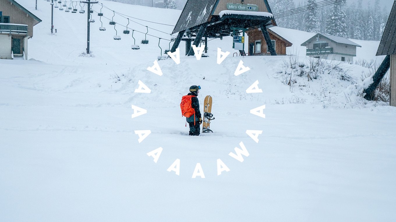 Snowboard in snow with board, AAAWA logo circle around snowy slopes and building behind him