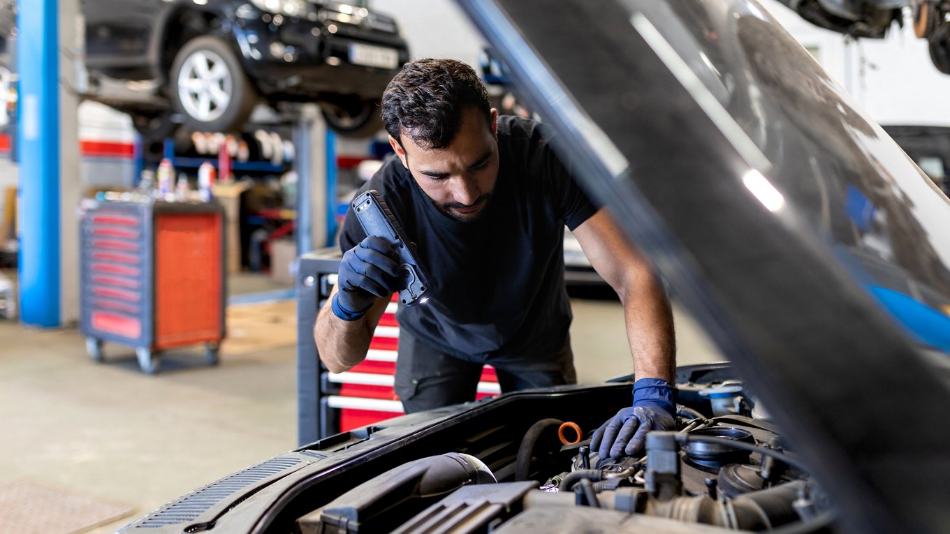 Serious male mechanic with flashlight examining lifted car while working in automobile service