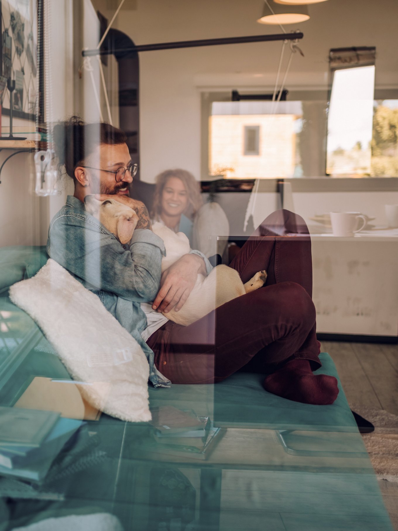 Beautiful happy family with dog talking and spending time together at home on a sofa. Couple petting their adopted puppy dog and relaxing at home on the weekend. Life with pets. Shot trough a window.
