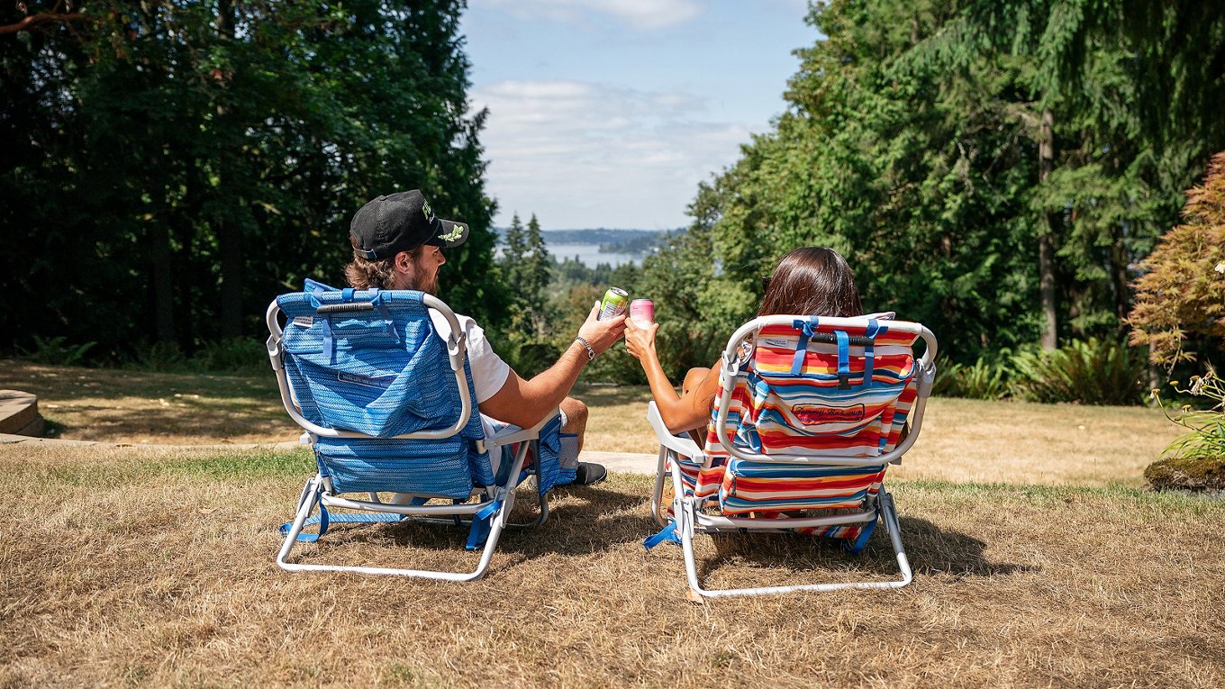couple sitting in color lawn chairs overlooking scenic pacific northwest area, holding our canned drinks to "cheers!"