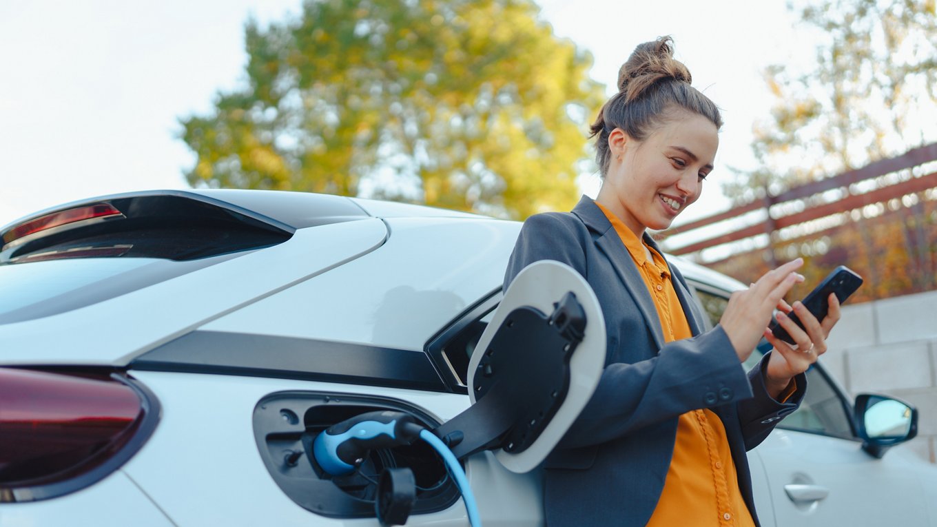 Young woman with smartphone waiting while electric car charging in home charging station, sustainable and economic transportation concept.