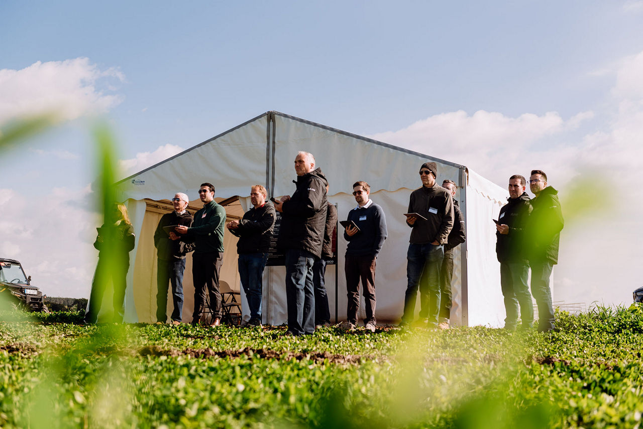 People wearing jackets, standing in front of a tent in a field, holding notepads, under a clear sky.-Alt-text description generated by Azure Vision AI model