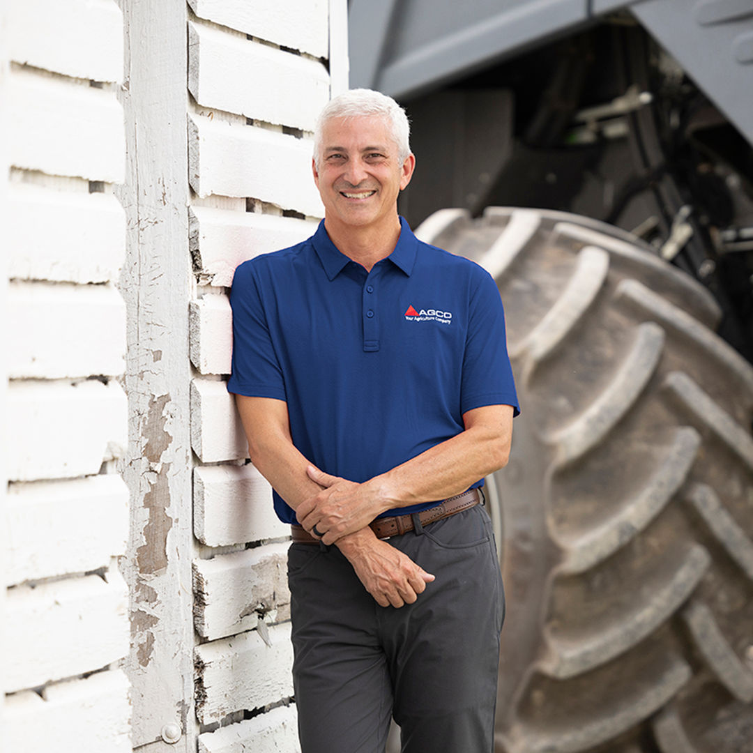 Smiling man in blue AGCO shirt standing against a weathered white wall, with a large tractor tire in background.-Alt-text description generated by Azure Vision AI model