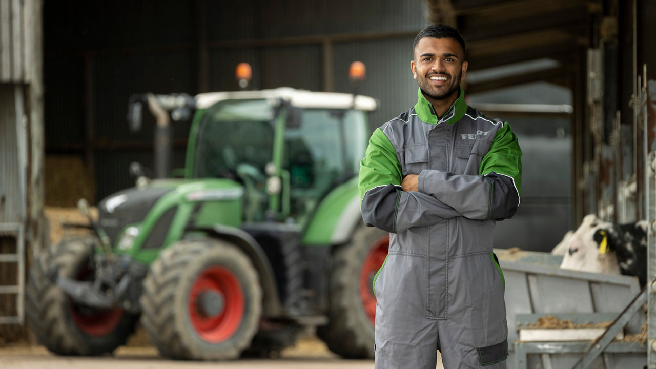 Man in Fendt workwear standing in foreground, arms crossed and smiling. Out of focus tractor in background