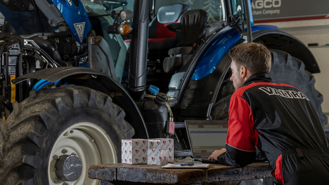 Man in red Valtra workwear looking at a blue Valtra tractor. There are parts boxes and an open laptop in front of him