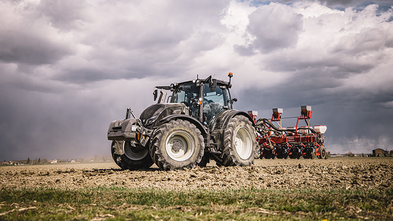 A Valtra N Series tractor is captured working in an open field. The setting is rural farmland with dramatic, overcast clouds dominating the sky. The scene emphasizes agricultural machinery in action, with earthy tones and a sense of productivity. 