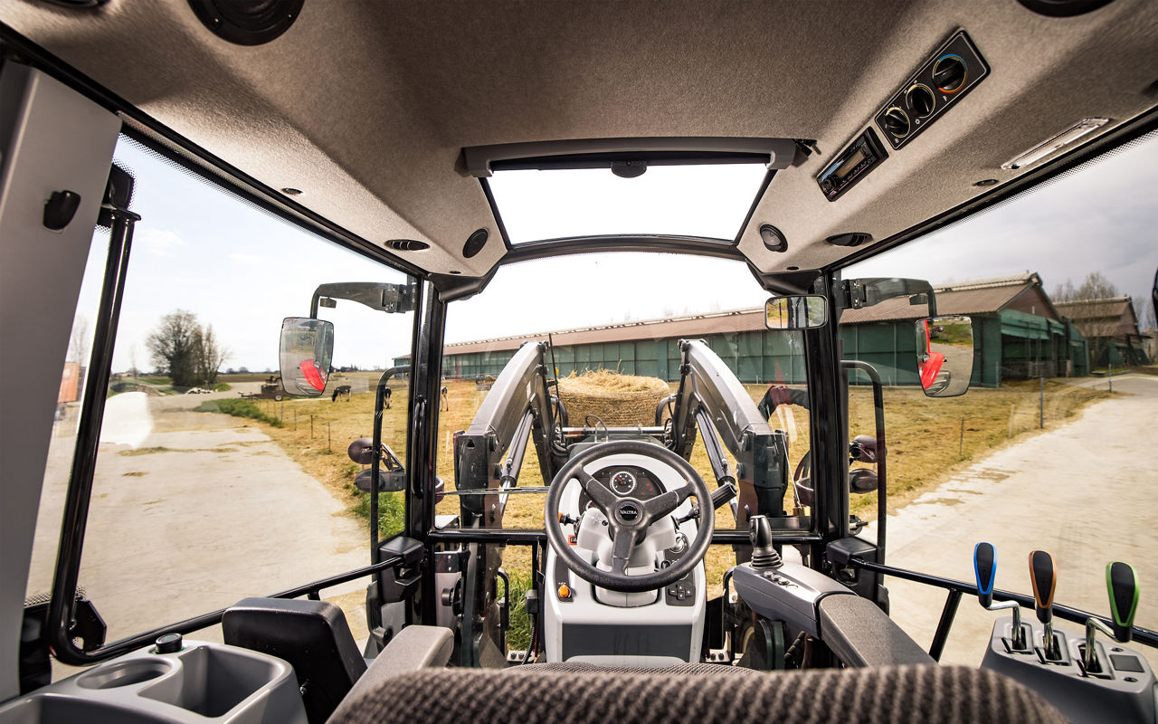 View from inside a modern tractor cab looking out over a farmyard. The image shows the steering wheel, control levers, and dashboard framed by large windows. Outside, farm buildings, equipment, and a dirt driveway are visible. The scene highlights agricultural machinery technology in a rural setting.