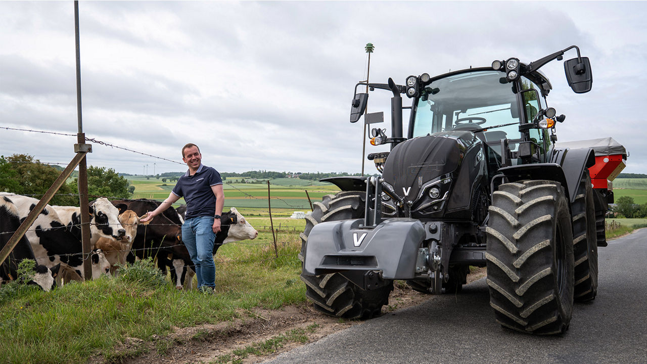 A man stands beside a group of cows near a fence, smiling at the camera. A black Valtra A Series tractor is parked on a rural road next to him. The scene is set in a green, open countryside under a cloudy sky, conveying a calm and productive agricultural environment.