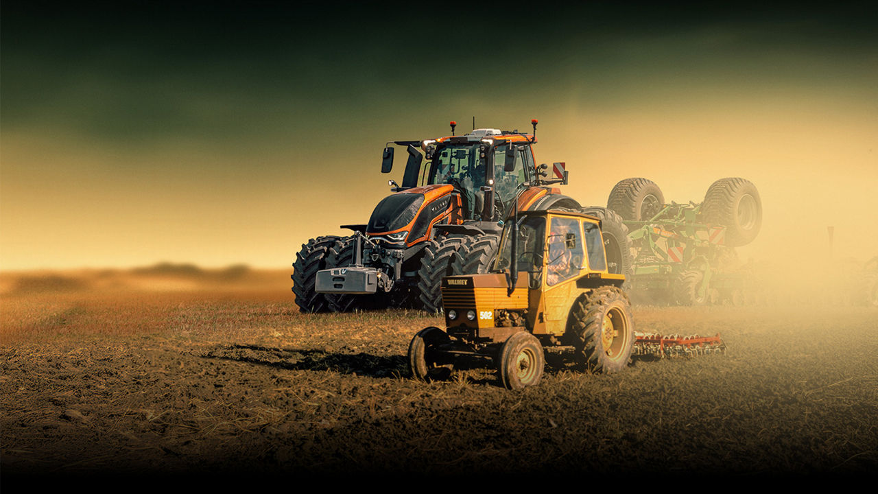 Two tractors stand on a wide, dusty field under a dramatic sky. A smaller yellow tractor is in the foreground pulling agricultural equipment. Behind it, a larger, more modern tractor looms with powerful tires and lights. The background fades into a hazy horizon, emphasizing the rural landscape and heavy machinery. The scene suggests active farm work and modern agriculture.