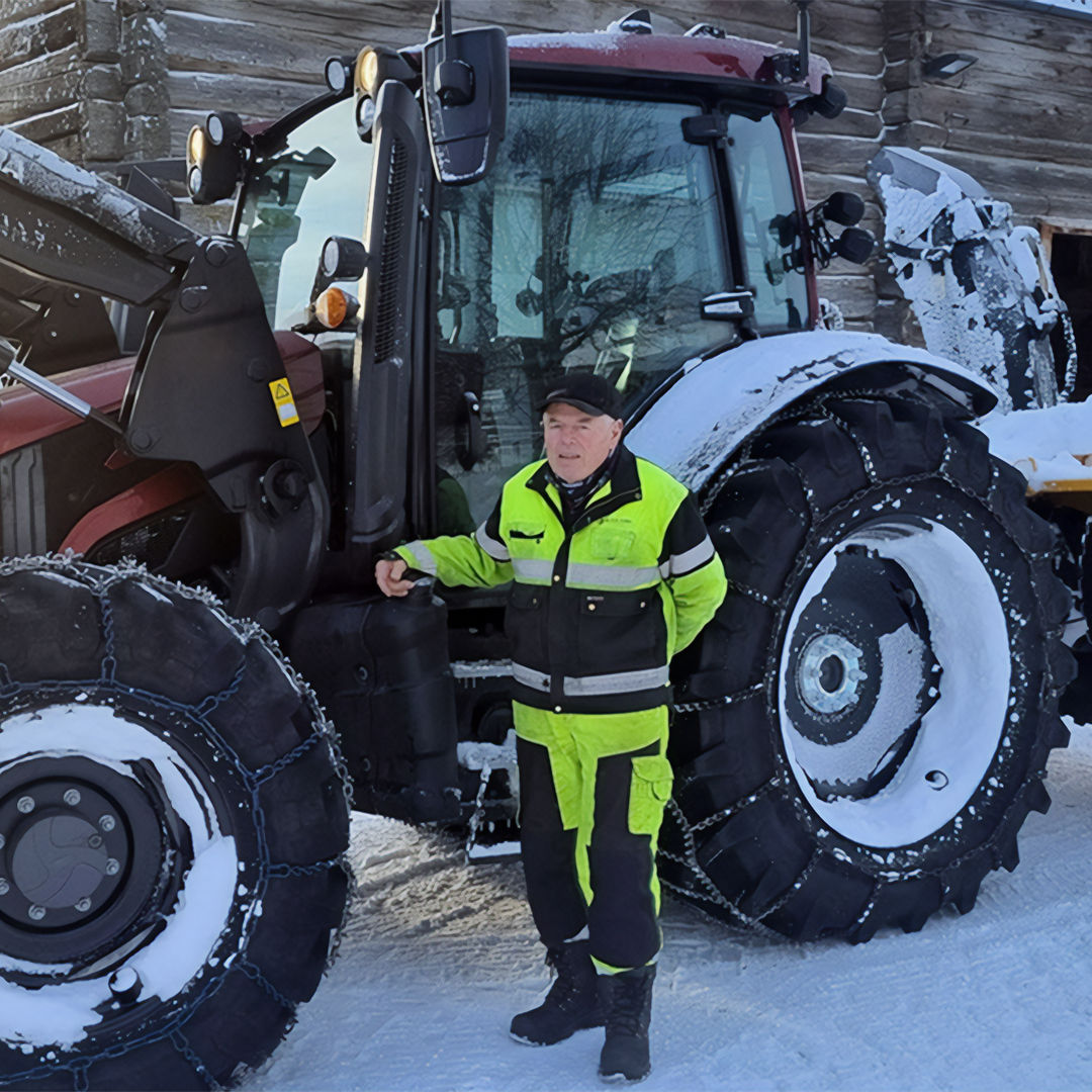 A person in high-visibility yellow and black workwear stands beside a Valtra G Series tractor in a snowy environment. The tractor is equipped with heavy-duty tires fitted with chains for traction on ice and snow. Snow covers parts of the vehicle and the ground, indicating cold winter conditions. A wooden building is visible in the background, adding a rural or industrial setting to the scene.