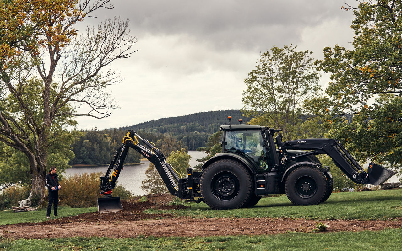 A Valtra tractor equipped with a front loader and a Hi-Tec 500F backhoe is operating on a landscaped area beside a lake. The backhoe is being used for excavation work while the front loader remains attached to the tractor. An operator is visible near the excavation area, with trees and water forming the background of the scene. The image focuses on the tractor and backhoe combination used for digging, earthmoving and specialist maintenance applications.