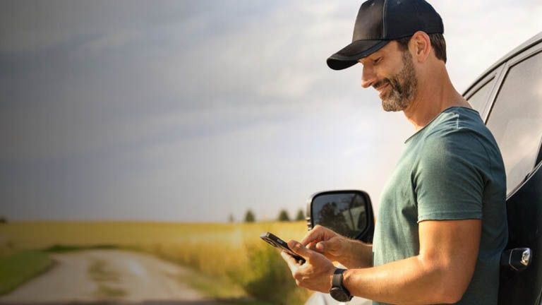 A man using a phone while smiling, standing by a wheat field