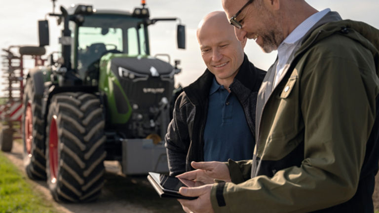 A farmer and a dealer looking at a phone and smiling with a tractor in the background