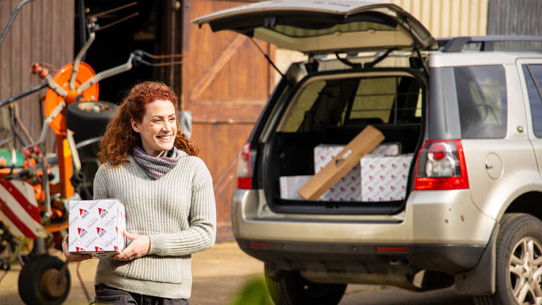 A woman holding an AGCO Parts box in front of a car with more boxes in the boot