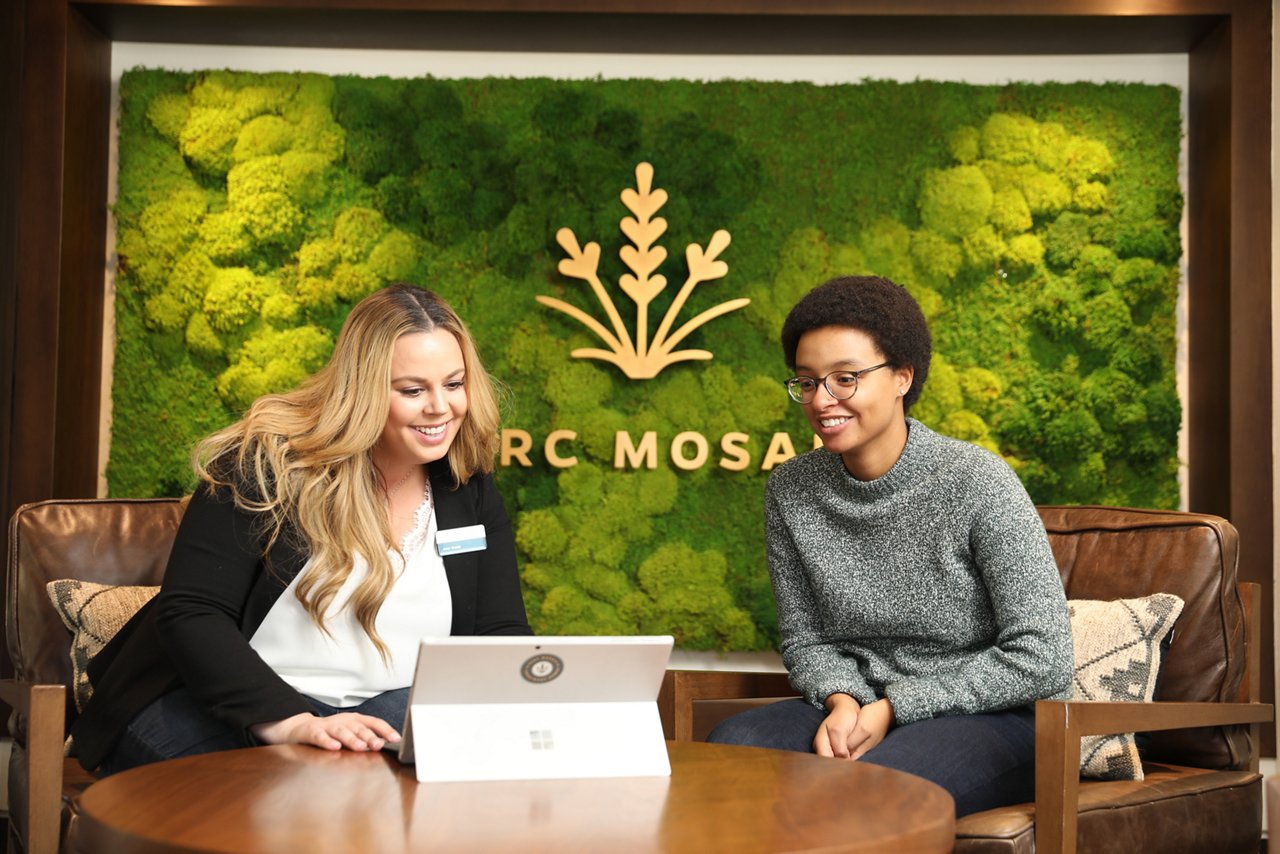 Two people at Parc Mosaic lobby looking at a computer screen. Green mossy wall in the background