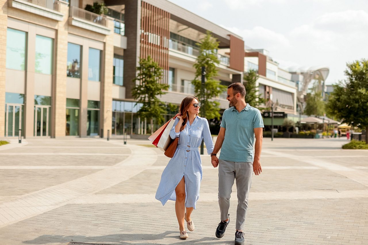 Two people stroll hand-in-hand through a vibrant shopping area, carrying bags and enjoying the warm sunshine on a delightful afternoon.