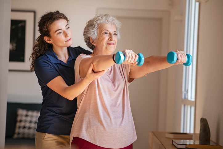 Old woman training with physiotherapist using dumbbells at home.