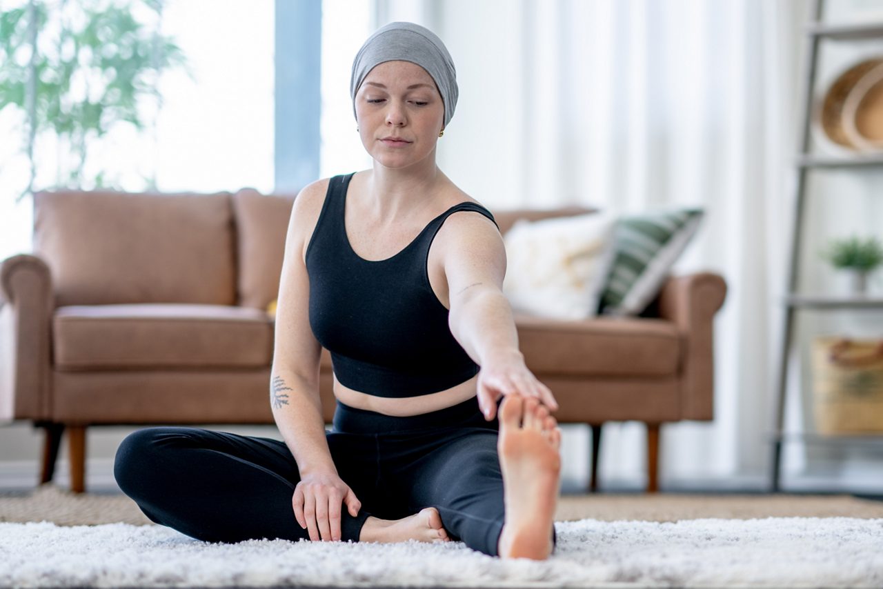 A young woman battling cancer sits on the floor in the comfort of her own home as she meditates.  She is dressed comfortably in athletic clothing and wearing a headscarf to keep warm.