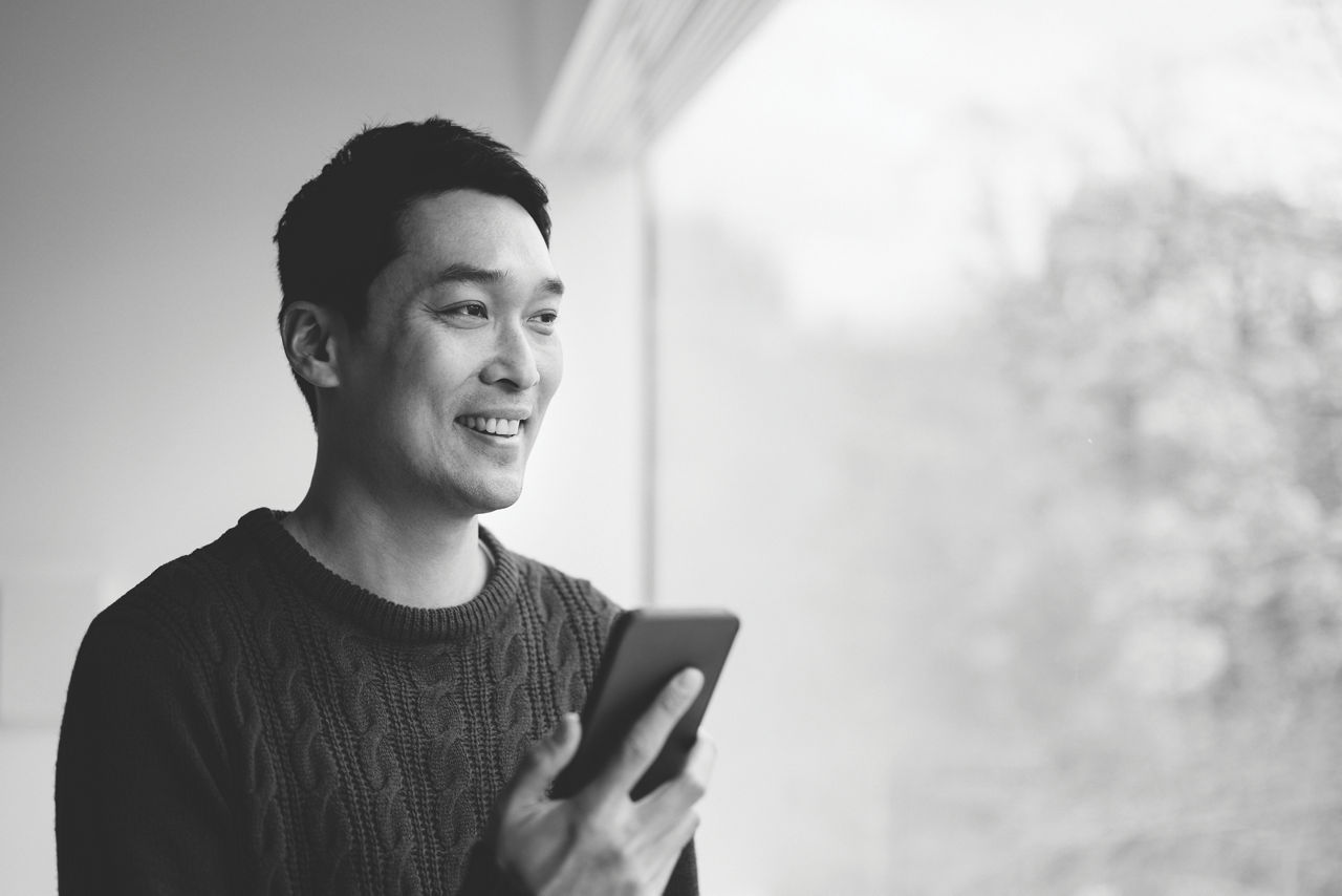 1317784222
Head and shoulders view of mature man with short black hair in pullover sweater holding mobile device and looking out window while talking on speaker phone.
