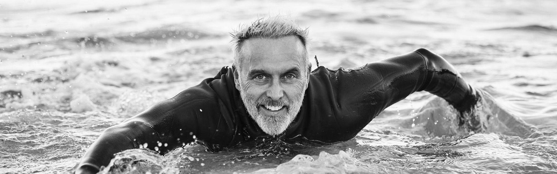 Man is at the beach during summer, in the ocean and surfing with his surfboard in a portrait. Mature, smile and surf in a wet suit for sport, fitness and health and happy in the sea water.