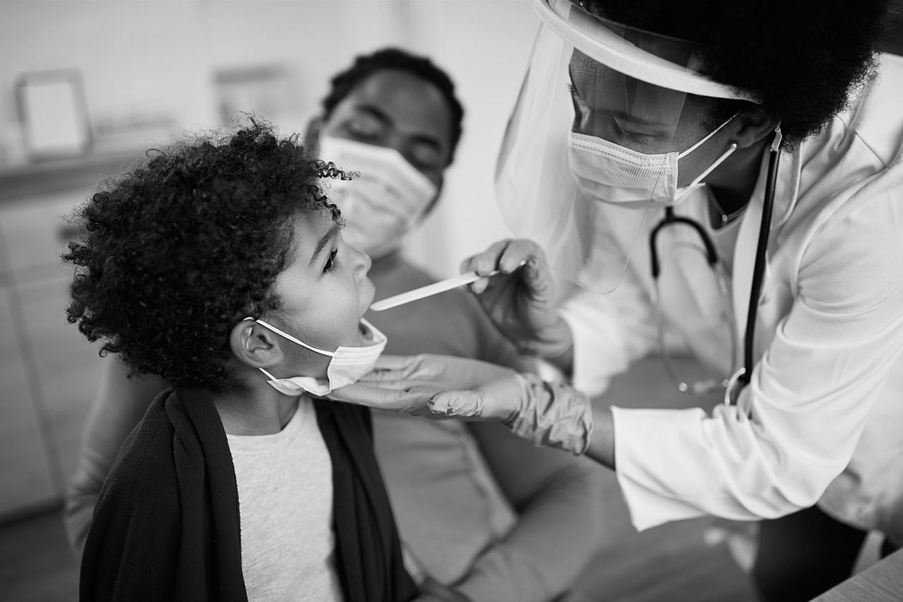 1287924870
Family doctor examining throat of a small black boy while visiting him at home during coronavirus pandemic.