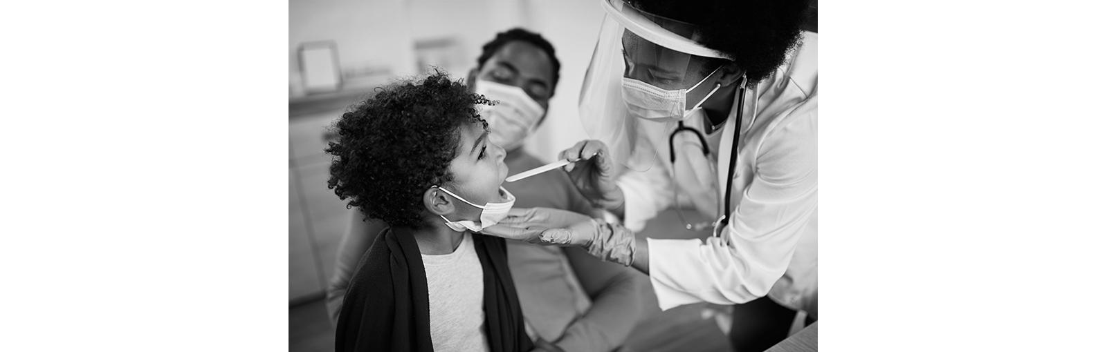 1287924870
Family doctor examining throat of a small black boy while visiting him at home during coronavirus pandemic.
