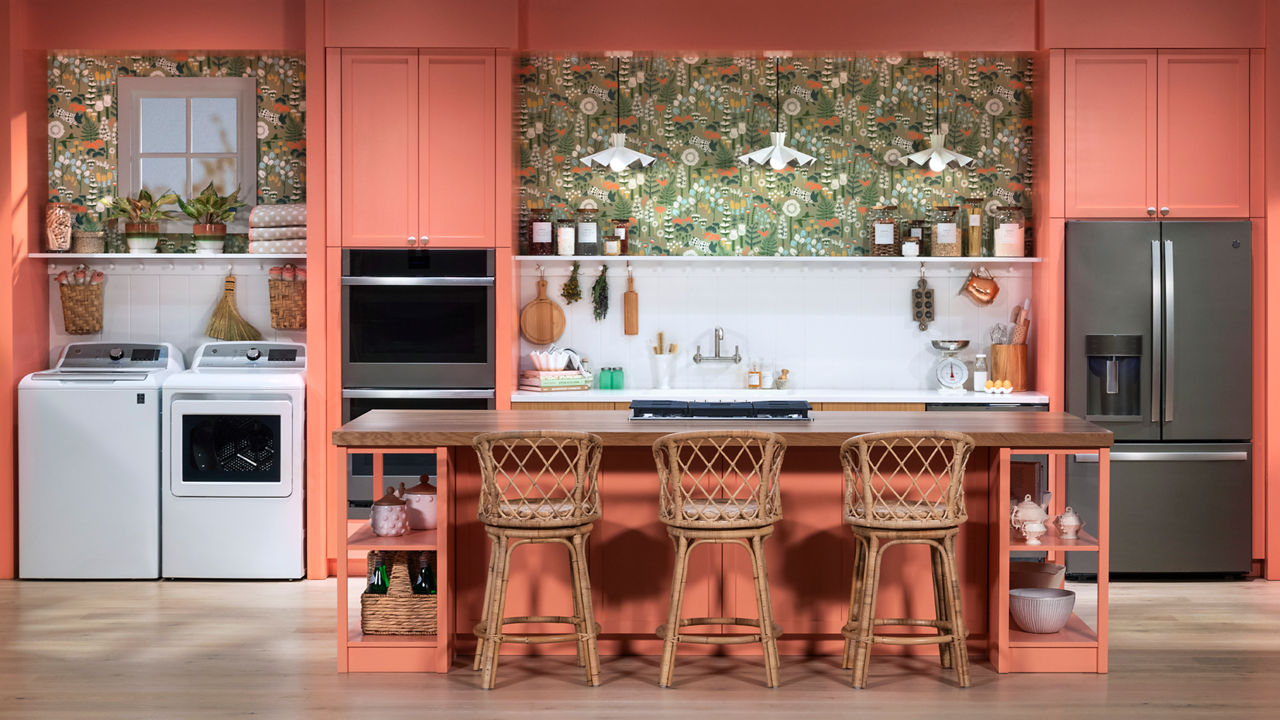 a peach colored kitchen with peach cabinets and island with three bar stools, bold patterned wall paper above a white quartz backsplash and countertops. 