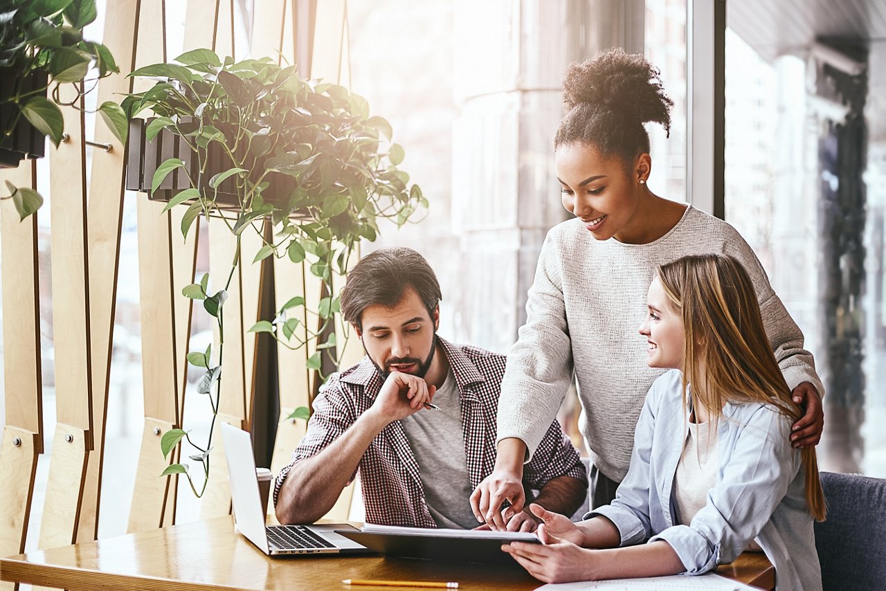 Three cheerful colleagues discussing new ideas in office. Bearded man looks on the chart, while women talk to him. A laptop computer, notebook and pencils are on the desk. Casual style. Concept of success