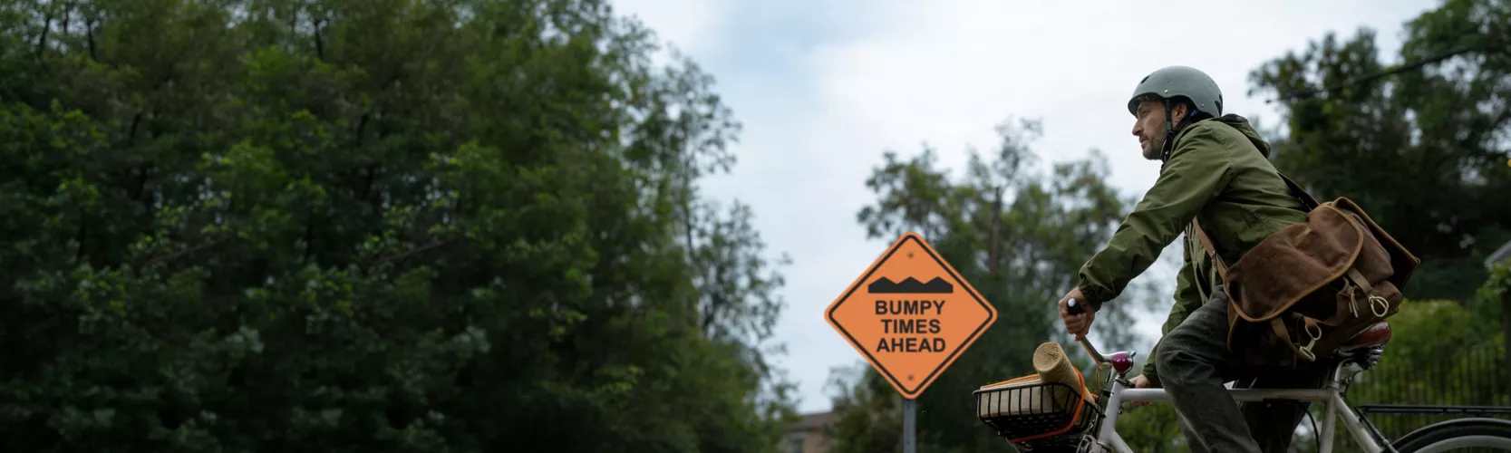 A person rides a bicycle along a road bordered by dense green trees. The cyclist wears a helmet and a green jacket, carrying a large brown shoulder bag and a basket on the bike. A bright orange diamond-shaped road sign reads "BUMPY TIMES AHEAD" with a wavy line graphic above the text. The sky is overcast, giving the scene a subdued, contemplative mood.