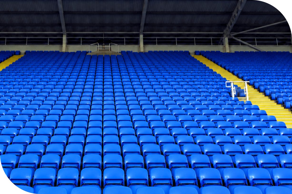 Rows of bright blue plastic seats fill an empty stadium stand. A central yellow stairway with white railings divides the seating sections and leads upward. The scene is covered by a dark metal roof structure, emphasizing the geometric pattern of the seats. The image conveys a quiet, anticipatory atmosphere before an event or game.